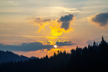 Gün batımında gökyüzünde renkli bulutlar, dağlık bir orman alanının arka planına karşı..