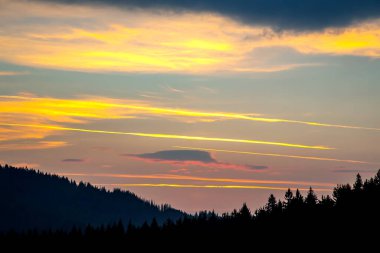 Gün batımında gökyüzünde renkli bulutlar, dağlık bir orman alanının arka planına karşı..