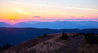 Gün batımında gökyüzünde renkli bulutlar, dağlık bir orman alanının arka planına karşı..