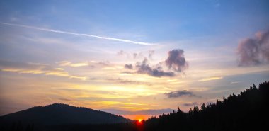 Gün batımında gökyüzünde renkli bulutlar, dağlık bir orman alanının arka planına karşı..