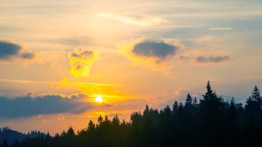 Gün batımında gökyüzünde renkli bulutlar, dağlık bir orman alanının arka planına karşı..