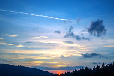 Gün batımında gökyüzünde renkli bulutlar, dağlık bir orman alanının arka planına karşı..