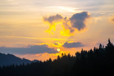 Gün batımında gökyüzünde renkli bulutlar, dağlık bir orman alanının arka planına karşı..