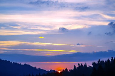 Gün batımında gökyüzünde renkli bulutlar, dağlık bir orman alanının arka planına karşı..