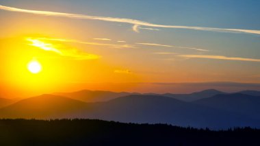 Gün batımında gökyüzünde renkli bulutlar, dağlık bir orman alanının arka planına karşı..