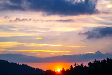 Gün batımında gökyüzünde renkli bulutlar, dağlık bir orman alanının arka planına karşı..