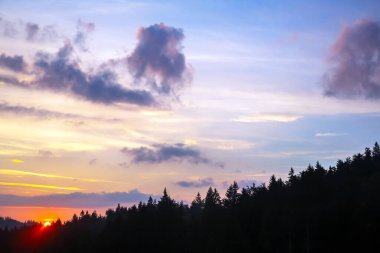 Gün batımında gökyüzünde renkli bulutlar, dağlık bir orman alanının arka planına karşı..