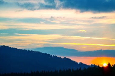 Gün batımında gökyüzünde renkli bulutlar, dağlık bir orman alanının arka planına karşı..