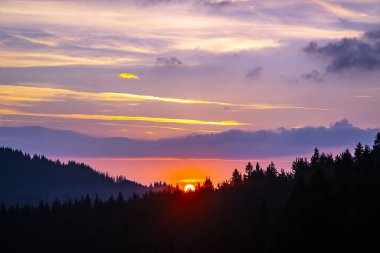 Gün batımında gökyüzünde renkli bulutlar, dağlık bir orman alanının arka planına karşı..