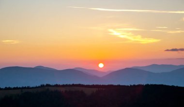 Gün batımında gökyüzünde renkli bulutlar, dağlık bir orman alanının arka planına karşı..