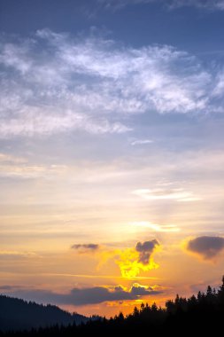 Gün batımında gökyüzünde renkli bulutlar, dağlık bir orman alanının arka planına karşı..