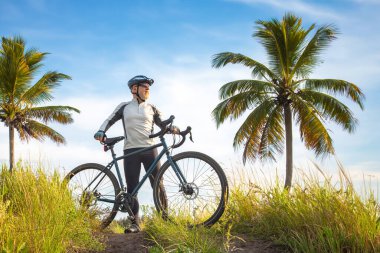 Bisikletli sarı elbiseli sakallı erkek bisikletçi doğadaki palmiye ağaçlarının arka planına karşı. Spor, hobiler ve sağlık için eğlence
