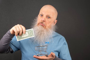 Büyük beyaz sakallı kel adam elinde bir dolar ve bir market sepeti modeli tutuyor. Finansal ekonomi konsepti. yoksulluk ve gıda krizi