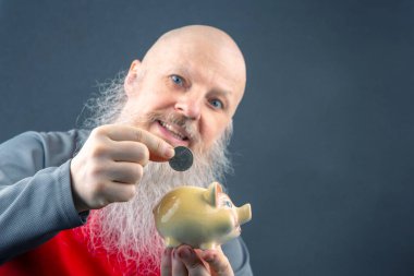 Uzun beyaz sakallı yaşlı bir adam kumbaraya bozuk para sokuyor. Finansal tasarruf, emeklilik planı ve kişisel finans yönetimi kavramı. Tarafsız bir arkaplanda gri ve kırmızı kıyafetler