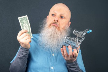 Büyük beyaz sakallı kel adam elinde bir dolar ve bir market sepeti modeli tutuyor. Finansal ekonomi konsepti. yoksulluk ve gıda krizi