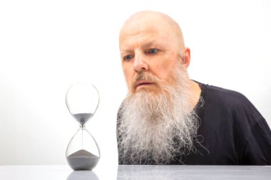 Beyaz sakallı kel bir adam zamanın sonsuz akışını simgeleyen kum saatini yakından inceliyor. Fotoğrafın minimalist tarzı, içgözlem duygusunu ve zamanın akışını arttırır.