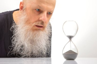 Büyük beyaz sakallı yetişkin bir adam pişmanlık ve endişeyle kum saatine bakar. Zamanın geçişi kavramı. Zaman geçiyor.