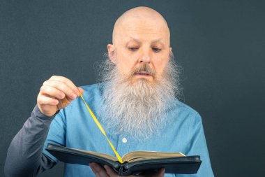 Seçkin gri sakallı bilge adam, kutsal kitabı okumaya dalmış, koyu gri bir arka planda, bağlılığı ve ruhani yansımayı sembolize ediyor.