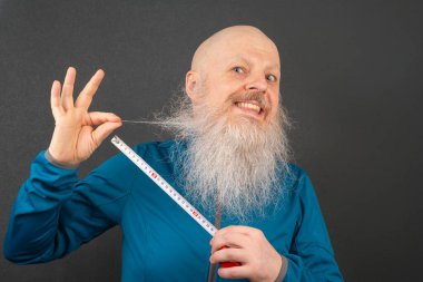 happy bald man demonstrating the length of his large white beard using a metric tape measure on a dark gray background clipart