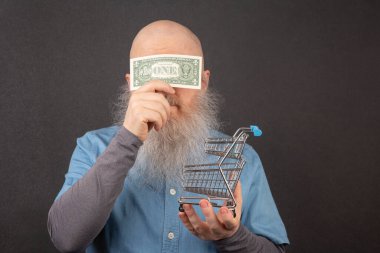 bald man with a large white beard holds a dollar and a model of a grocery basket in his hands. financial economics concept. poverty and food crisis clipart