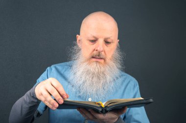 Yaşlı adam, gri sakallı, koyu gri arka planda oturmuş, inanç ve bilgeliğin altını çizerek İncil 'den kutsal kitabı tutkuyla okuyor.