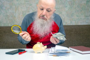 Ciddi bir görünümü olan yaşlı bir adam büyüteçle dolar banknotlarını teftiş ediyor, bir domuz kumbarası, akıllı telefon ve bir yığın faturayla bir masada oturuyor, yaşlılıkta mali kaygıları temsil ediyor.