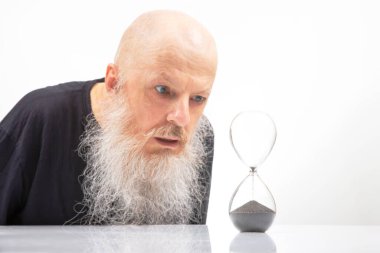 Beyaz sakallı kel bir adam zamanın sonsuz akışını simgeleyen kum saatini yakından inceliyor. Fotoğrafın minimalist tarzı, içgözlem duygusunu ve zamanın akışını arttırır.