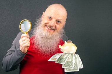 Sakallı kıdemli adam büyüteç ve para dolu domuz kumbarası gösteriyor, kişisel finans, emlak tasarrufu, ev sahipliği planlama ve yatırım stratejilerinin temaları vurgulanıyor