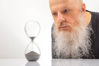 Büyük beyaz sakallı yetişkin bir adam kum saatindeki düşen kum tanelerine pişmanlık ve endişeyle bakar. Zamanın geçişi konsepti. Zamanın geçişi.