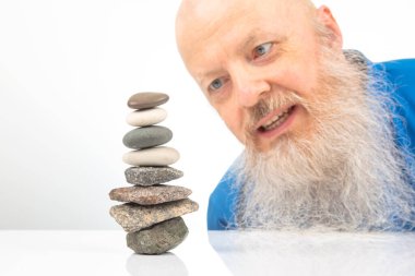 Hevesli Adam 'dan Bilinçli Kaya Dengeleme Hareketi. Mutlu sakallı adam beyaz arka planda taştan kuleye bakıyor. Hayattaki denge kavramı