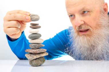 Neşeli sakallı adam dikkatli bir şekilde pürüzsüz kayaları dengeli bir kuleye yığıyor, sakin bir ortamda sabır ve odaklanma gösteriyor, doğal taş düzeninde farkındalığı ve yaratıcılığı vurguluyor.