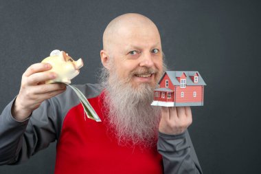 Uzun gri sakallı kel bir adam, elinde bir dolarlık banknot ve küçük bir model evi olan domuz kumbarası tutuyor. Tasarruf ve gayrimenkul yatırımlarını temsil ediyor.