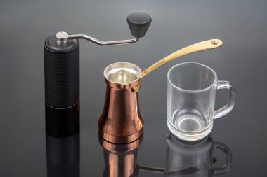 Metalik saplı siyah manuel kahve öğütücüsü, altın saplı bakır bir çakmak ve parlak siyah bir yüzey üzerinde cam bardak, kahve demleme seansları için ideal.