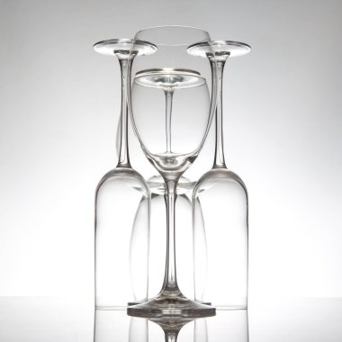 Narin gövdeli kristal berrak şarap kadehleri yan yana diziliyor. Yansımaları, nötr gri bir arkaplana karşı parlak bir yüzey üzerindeki kompozisyonu artırıyor..