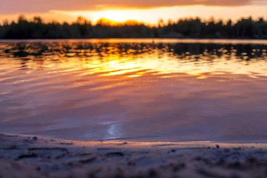 Huzurlu bir göl üzerinde sersemletici bir gün batımı, dalgalı sulara canlı turuncu renkler yansıtıyor, bir orman silueti tarafından çerçevelenmiş. Doğa ve manzara fotoğrafçılığı için ideal..