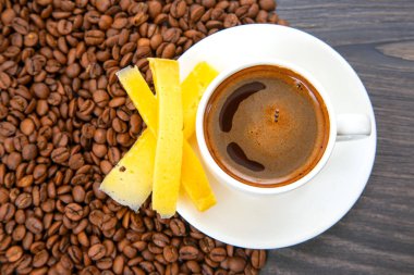 Beyaz bir fincan espresso, üzerinde sarı peynir çubukları olan bir tabağın üzerinde oturur. Yukarıdan yakalanmış, koyu ahşap bir masada kavrulmuş kahve çekirdekleri ile çevrilidir..