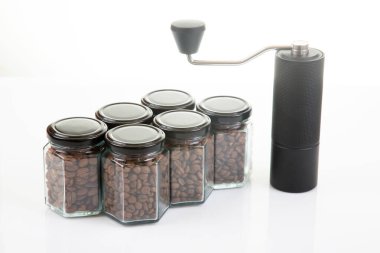 Kahve çekirdekleri için siyah manuel kahve öğütücüsü ve siyah kahve çekirdekleri ile dolu altı bardak şeffaf kavanoz ve beyaz arka planda kahve demlemek için toprak takımı..