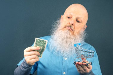 Büyük beyaz sakallı kel adam elinde bir dolar ve bir market sepeti modeli tutuyor. Finansal ekonomi konsepti. yoksulluk ve gıda krizi