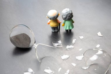 Minyatür yaşlı bir çift, zamanın geçişini ve kırılganlığını simgeleyen karanlık bir yüzeye dökülen kumla kırık bir kum saatinin yanında duruyor..