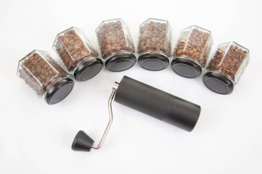 Kahve çekirdekleri için siyah manuel kahve öğütücüsü ve siyah kahve çekirdekleri ile dolu altı bardak şeffaf kavanoz ve beyaz arka planda kahve demlemek için toprak takımı..