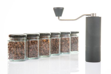 Kahve çekirdekleri için siyah manuel kahve öğütücüsü ve siyah kahve çekirdekleri ile dolu altı bardak şeffaf kavanoz ve beyaz arka planda kahve demlemek için toprak takımı. Kahve için mutfak aletleri..