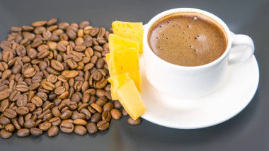 Beyaz espresso bardağının yakın çekimi, peynir çubukları, ve kahve çekirdekleri karanlık bir masada, basit ama zarif bir yemek eşleşmesi sergiliyor..