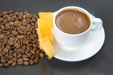 Kavrulmuş kahve çekirdekleri koyu bir yüzeyde beyaz bir fincan espresso ve sarı peynir çubuklarını sarıp sıcak ve davetkar bir kompozisyon oluşturur..