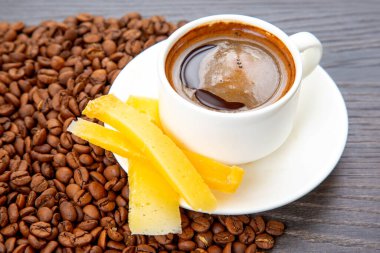 Beyaz bardakta dumanı tüten bir espressonun üst görüntüsü, yanında sarı peynir çubukları ve koyu ahşap arka planda dağınık kahve çekirdekleri var..