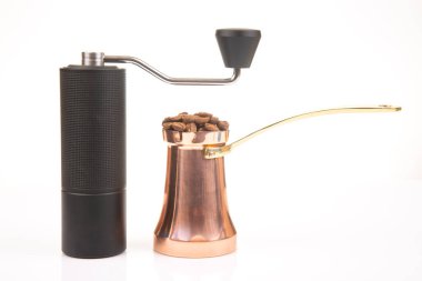 Siyah kahve öğütücüsü, bakır bir kahve ve beyaz arka planda kavrulmuş kahve çekirdekleri, geleneksel kahve yapımının özünü yakalıyor..