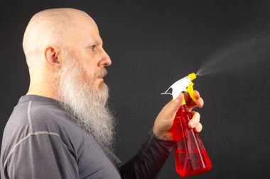 Kel ve sakallı bir adam koyu gri arka planda sprey şişesinden sıvı sıçratıyor.