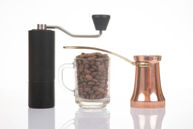 Sade bir kahve öğütücüsünün yakın çekimi, parlak bir bakır çakmak, ve kahve çekirdekli cam bardak, kahve hazırlama görselleri için temiz beyaz bir zemin üzerine kurulmuş.