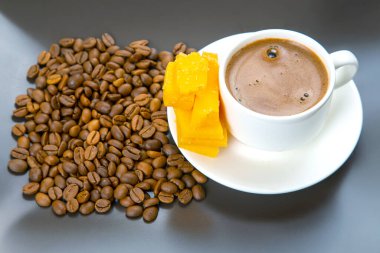 Kavrulmuş kahve çekirdekleri koyu bir yüzeyde beyaz bir fincan espresso ve sarı peynir çubuklarını sarıp sıcak ve davetkar bir kompozisyon oluşturur..