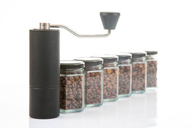 Kahve çekirdekleri için siyah manuel kahve öğütücüsü ve siyah kahve çekirdekleri ile dolu altı bardak şeffaf kavanoz ve beyaz arka planda kahve demlemek için toprak takımı..