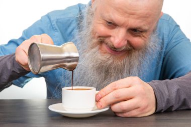 Uzun beyaz sakallı mutlu bir adam ev yapımı kahvaltı atmosferinde beyaz bir bardağa kahve dolduruyor. sıcak içecek.